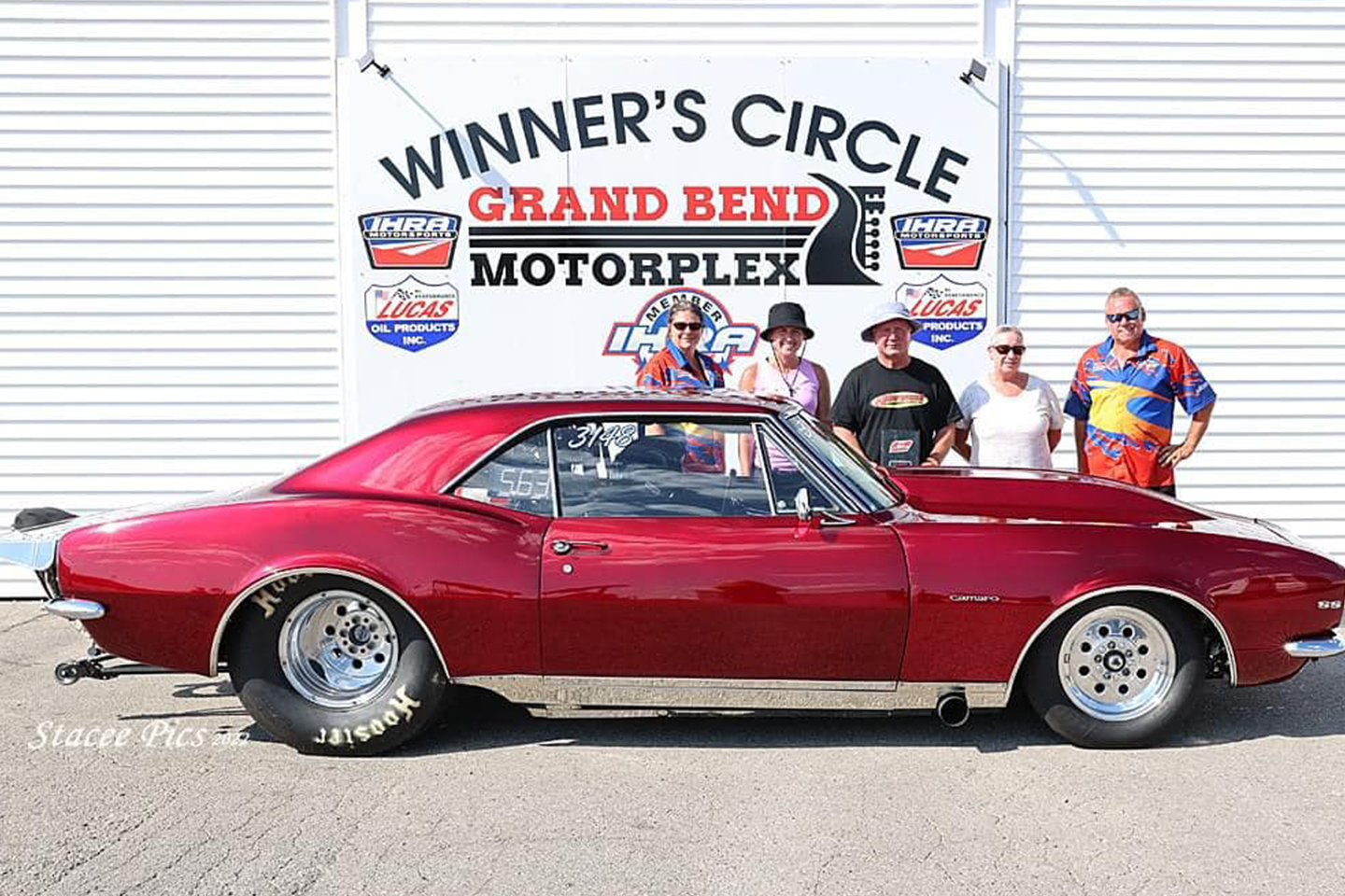 A maroon classic car, parked in front of the Winner’s Circle sign at Grand Bend Motorplex, surrounded by four people posing for a celebratory photo.