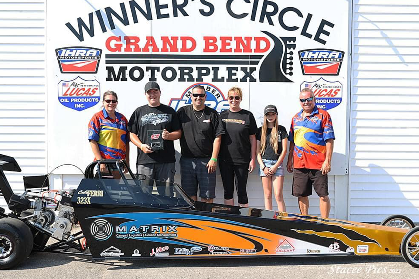 A group of six people standing in front of the Winner’s Circle at Grand Bend Motorplex, posing with a black and orange dragster and holding a trophy.
