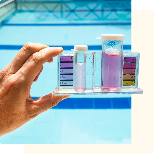 A hand holding a swimming pool water test kit with pink-colored chemical reagents, shown against the backdrop of a bright blue pool.