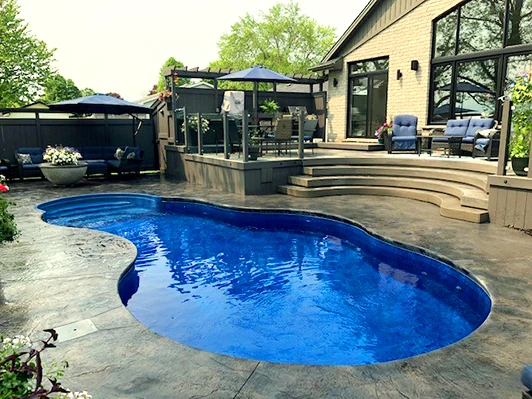 A freeform in-ground swimming pool with a bright blue interior, surrounded by a stamped concrete patio. A multi-level deck with umbrellas, outdoor seating, and a brick house stand in the background, framed by lush greenery.
