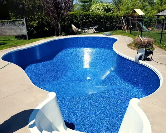 A freeform in-ground swimming pool with a newly installed bright blue liner, surrounded by a concrete patio. A hammock, children’s play set, and a fence line the backyard in the background.