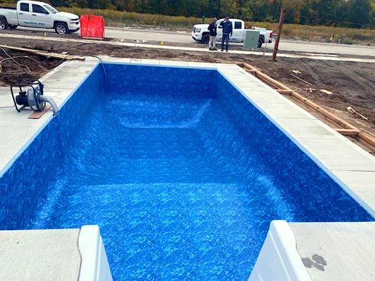 A rectangular in-ground swimming pool with a newly installed bright blue liner, surrounded by fresh concrete. Construction materials, vehicles, and workers are visible in the background.