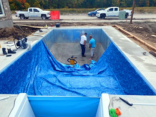 A rectangular in-ground swimming pool under construction with a partially installed blue liner. Two men stand inside the pool, working on the interior. Construction vehicles and materials are visible in the background.