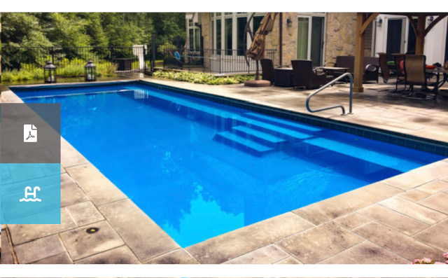 A rectangular, in-ground swimming pool with bright blue water, stone paver decking, and a metal handrail. A stone house and patio furniture are visible in the background, surrounded by greenery.