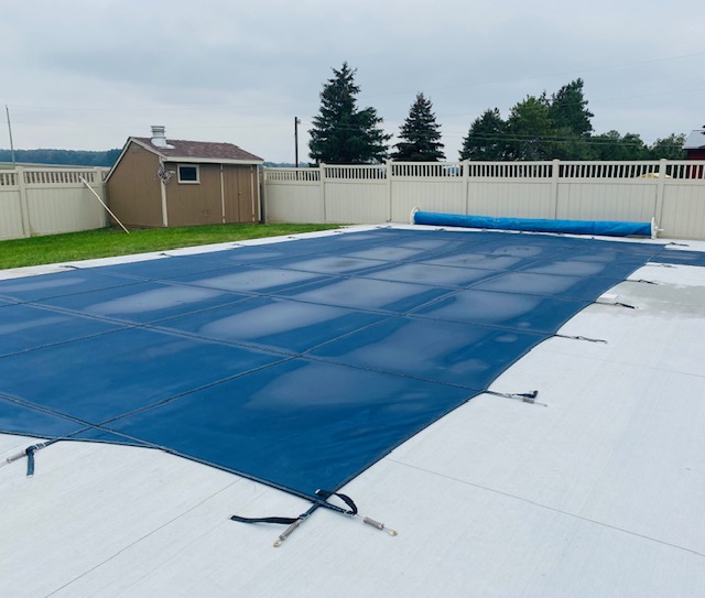 A rectangular in-ground pool covered with a solid blue cover, set in a concrete patio area and enclosed by a beige fence, with a small shed and trees in the background under an overcast sky.