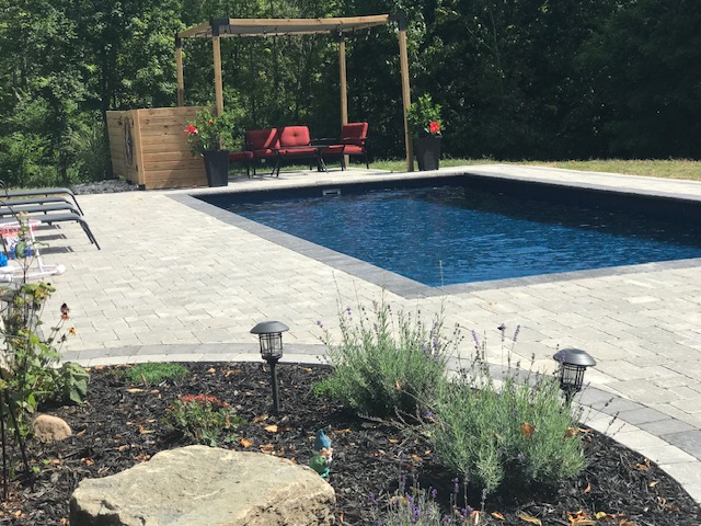 A rectangular, in-ground swimming pool with a dark blue interior, surrounded by a stone paver patio. In the background, there’s a pergola with red cushioned seating and lush greenery, while landscaped flowerbeds and trees complete the backyard setting.