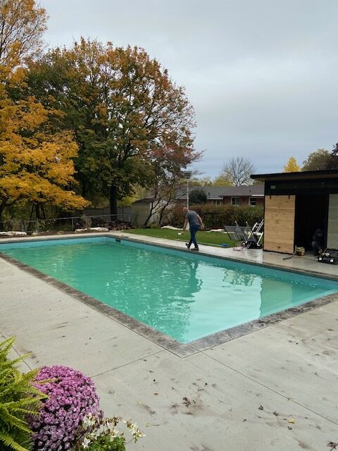 A rectangular, in-ground swimming pool in a backyard surrounded by autumn foliage, with a person walking along the pool’s edge and a small modern outbuilding to the right.