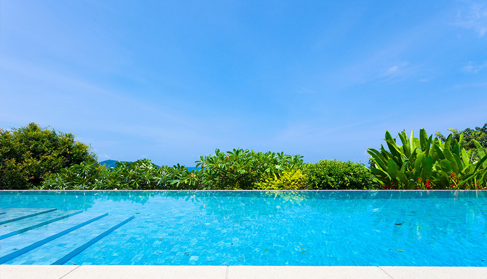 A bright, clear swimming pool in the foreground, bordered by lush green tropical foliage, with a wide blue sky stretching overhead.