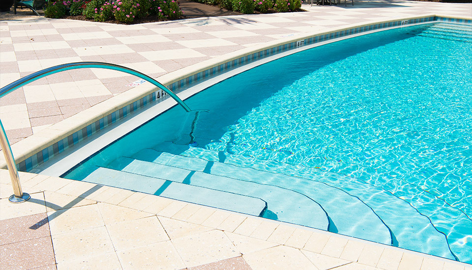 bright, outdoor swimming pool with clear turquoise water, featuring steps and a curved metal handrail, surrounded by a checkered tile deck.